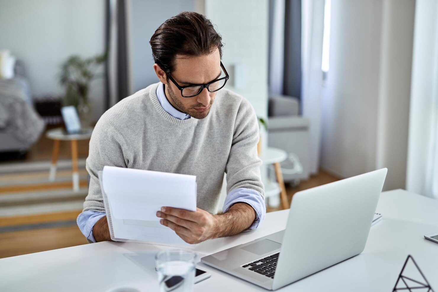 A man seated at a desk with a laptop open holding a note pad and glancing at the screen.